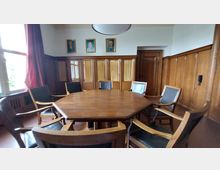 Holzvertäfelter Besprechungsraum mit einem achteckigen Holztisch und umliegenden gepolsterten Holzstühlen. An der Wand befinden sich gerahmte Porträts sowie hohe Schränke mit Glastüren und Vorhängen, daneben eine Holztür; links ein großes Fenster mit roten Vorhängen.