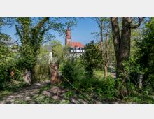 Blick auf eine Kirche mit rotem Dach und hohem Turm, umgeben von dicht bewachsenen Bäumen und Sträuchern. Im Vordergrund steht ein gemauerter Torbogen mit geschlossenem Metallgitter, eingebettet in eine grüne Parklandschaft.