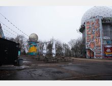 Offener Außenbereich mit Bierbänken und geschlossenen Sonnenschirmen auf einem unbefestigten Platz, umgeben von kahlen Bäumen. Rechts steht ein hoher, zylindrischer Bau mit großer geodätischer Kuppel und Graffiti-Fassade, links ein kleinerer Turm mit Kugelaufsatz und bunter Bemalung; über dem Platz hängen Lichterketten.