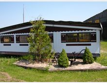 Ein länglicher, weiß-brauner Caravan mit rechteckigen Fenstern steht auf einer Wiese. Vor dem Caravan befindet sich ein kleines Gartenbeet mit einem Baum, zwei kleinen Tannen und einem dekorativen Pflug.