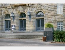 Steinerne Fassade eines historischen Gebäudes mit drei großen Rundbögen, verziert mit Reliefs und dekorativen Elementen. Vor den Bögen befinden sich vergitterte Eingangstüren, eine Treppe führt zum Eingang, und im Vordergrund stehen ein Pflanzenkübel und ein Mülleimer.