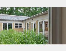 Ein Innenhof mit bepflanzten Grünflächen, umgeben von einem einstöckigen Gebäude mit gelben Backsteinwänden und weißen Fenstern. Im Hintergrund sind hohe Bäume zu sehen, die den Hof teilweise umrahmen.