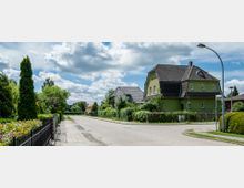 Straßenszene in einer Wohngegend mit einem grünen Einfamilienhaus und gepflegten Hecken und Gärten. Die Straße ist gepflastert und von Laternen sowie Bäumen und Sträuchern gesäumt, mit einem wolkigen Himmel im Hintergrund.