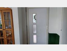 Innenflur mit einer weißen Eingangstür, die drei schmale, übereinander angeordnete Sichtfenster besitzt. Links steht eine hohe Vitrine aus Holz mit Glastüren, rechts ist ein grün gestrichener Wandbereich zu sehen.