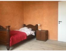 Ein schlicht eingerichtetes Schlafzimmer mit einer orangefarbenen Wand, einem Einzelbett aus dunklem Holz mit Schnitzverzierungen, einem roten Bettüberwurf und einem kleinen Holznachttisch mit einer weißen Lampe. Der Boden besteht aus hellem Holz, und rechts ist eine weiße Tür sichtbar.