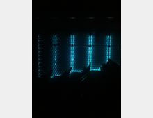 Dunkler Innenraum mit einer metallischen Wand, die von leuchtenden blauen LED-Streifen erhellt wird. Die Wand weist ein geometrisches, texturiertes Muster auf, im Vordergrund sind schemenhaft Sitzplätze erkennbar.