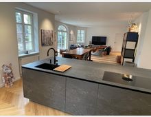 Moderne Wohnküche mit offenem Wohnbereich. Im Vordergrund eine Kücheninsel mit grauer Arbeitsplatte und integriertem Waschbecken, im Hintergrund ein Essbereich mit Holzfußboden, großen Fenstern und einem Wohnzimmer mit Sofa und Flachbildfernseher.