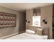 Schlafzimmer mit beiger Wandfarbe, einem Bett mit karierter Polsterkopfstütze und weißer Bettwäsche. An der Wand hängt ein Kunstwerk mit Campbell's Suppendosen, und rechts über dem Bett ist ein beleuchtetes Wandregal mit Büchern und Dekoration integriert.