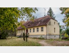 Ein gelb gestrichenes, einstöckiges Gebäude mit rotem Ziegeldach und weiß gerahmten Fenstern, das auf einem Sockel aus Feldsteinen steht. Umgeben von Bäumen und Grünflächen, mit einem gepflasterten Weg, der zur Eingangstreppe führt.