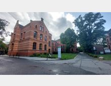 Backsteingebäude im Stil der Neogotik, umgeben von einer Grünfläche mit Bäumen und Gehwegen. Im Vordergrund stehen ein Informationstafel und ein Fahrrad, im Hintergrund lichtet sich der wolkenbedeckte Himmel.