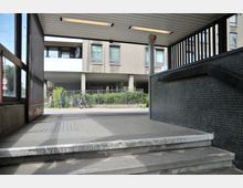 Eingangsbereich eines Wohngebäudes mit Treppen und einer Rampe, flankiert von dunklen Wandfliesen. Im Hintergrund sind ein moderner Balkon, Fenster und Fahrräder vor einem Zaun und grünen Pflanzen zu sehen.