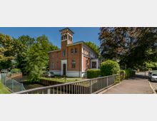 Backsteinhaus mit Turm in klassizistischer Architektur, umgeben von Bäumen, einer kleinen Brücke und einem angrenzenden Wasserlauf; im Vordergrund befindet sich ein Gehweg.