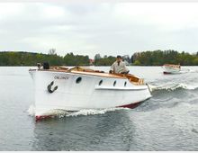 Ein weißes Motorboot mit der Aufschrift „OBLOMOV“ fährt auf einem ruhigen Gewässer, gesteuert von einer Person in einem Poncho und Mütze. Im Hintergrund ist ein weiteres ähnliches Boot und eine Landschaft mit Bäumen sowie Gebäuden am Ufer zu sehen.