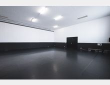 Großer, leerer Raum mit weiß gestrichenen Wänden und schwarzem Bodenbelag. Die Wände haben eine schwarze Sockelleiste, es gibt eine schwarze Tür an einer Wand, und die Decke ist mit Rasterplatten bedeckt sowie von mehreren Deckenleuchten beleuchtet.