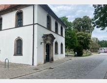 Historisches zweistöckiges Gebäude mit weißer Fassade und hohen, bogenförmigen Fenstern, umgeben von einer gepflasterten Fläche. Im Hintergrund sind Bäume und Grünflächen zu sehen, daneben ein Container mit roten und weißen Elementen.