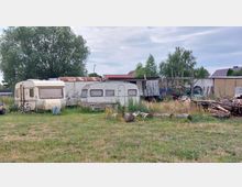 Eine Wiese mit zwei geparkten Wohnwagen, einem Schuppen mit Wellblechdach und gestapeltem Brennholz. Im Hintergrund stehen große Bäume und einige Häuser, umgeben von verschiedenen Gegenständen wie Fahrrädern und Werkzeugen.