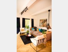 Modernes Wohnzimmer mit Holzparkettboden, einer großen beigen Eckcouch, farbigen Kissen, einem schwarzen Couchtisch und weißen Sesseln mit Metallrahmen. Große Fenster mit dunkelblauen Vorhängen, Pflanzen und abstrakte Wandkunst schaffen eine gemütliche Atmosphäre.