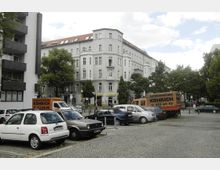 Straßenszene in einer Wohngegend mit mehrstöckigen, historischen Wohnhäusern aus hellem Putz im Hintergrund. Im Vordergrund ein Parkplatz mit parkenden Autos und einem großen Transporter mit Werbung für eine Autovermietung, umgeben von Bäumen und gepflasterten Gehwegen.
