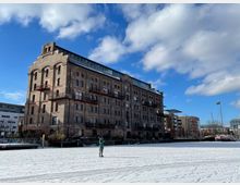 Backsteingebäude mit mehreren Etagen und modernen Balkonen an einer Hafenumgebung, flankiert von Wohnhäusern und Hafenanlagen. Im Vordergrund eine schneebedeckte Fläche, darüber ein blauer Himmel mit Wolken.