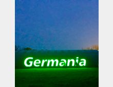 Eine nächtliche Szene zeigt eine große, grün leuchtende Beschriftung mit dem Wort "Germania" vor einem dunklen, gewölbten Gebäude. Der Untergrund ist eine grasbewachsene Fläche, im Hintergrund sind schwach erkennbare Bäume zu sehen.