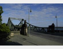 Eine grüne Stahlbrücke mit geschwungenen Bögen und dekorativen Statuen aus Stein an den Brückenköpfen, die über einen Fluss führt. Im Vordergrund eine gepflasterte Fläche mit Laternen und Bäumen am linken Bildrand.