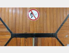 Holztür mit senkrechten Holzlatten, verstärkt durch schwarze Metallbeschläge. In der Mitte befindet sich ein rundes Verbotsschild mit einer durchgestrichenen Flamme und einem Streichholz, das auf Feuergefahr hinweist.