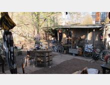 Ein kleiner, rustikaler Hinterhof mit diversen Gegenständen wie Fahrrädern, einer Feuerstelle, Holzmöbeln und Werkzeugen. Im Hintergrund steht ein einfaches Gebäude mit Wellblechdach, umgeben von Bäumen und Gebrauchsgegenständen.