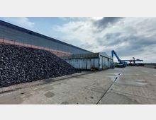 Industriegelände mit einer großen Halle, einem Stapel von schwarzen Materialien (möglicherweise Schrott) und mehreren Krananlagen im Hintergrund. Im Vordergrund ein betonierter Platz mit leicht wolkigem Himmel.