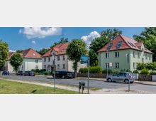 Straßenszene in einer Wohngegend mit mehreren zweistöckigen Häusern mit rotem Satteldach und hellgrüner Fassade; am Straßenrand parkende Autos und Straßenschilder für "Schloßstraße" und "Mozartstraße". Im Hintergrund stehen Bäume, und der Himmel ist blau mit vereinzelten Wolken.