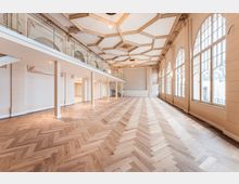 Großer, leerer Saal mit hellem Fischgrätparkett, hoher kassettierter Stuckdecke und umlaufender Empore mit Glasgeländer. Rechts sorgen hohe, bogenförmige Fensterfronten für viel Tageslicht; im Hintergrund führt eine Treppe zur oberen Ebene.