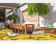 Sonnige Terrasse mit Holzboden, auf der sich ein langer Esstisch aus Holz mit geflochtenen Stühlen sowie eine Sitzgruppe mit weißen Polstern und grünen Kissen befindet; im Hintergrund eine Wand mit antik wirkendem Wandbild und einem kleinen Baum. Links ist eine Glas-Schiebetür zu einem Innenraum sichtbar.