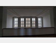 Ein heller Innenraum mit einer Wand aus fünf großen, rechteckigen Fenstern, die in schmalen weißen Rahmen gefasst sind. Die Fensterfront wird von schlichtem, hellem Putz umgeben, und eine niedrige Brüstung aus Holz verläuft entlang der unteren Fensterkante.