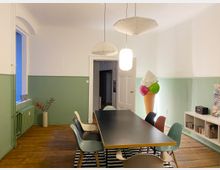 Ein heller Raum mit Holzboden, pastellgrünen Wänden und einer großen schwarzen Tischplatte, umgeben von bunten Stühlen. Im Hintergrund sind ein Türdurchgang, eine leuchtende Eistüten-Dekoration und ein Regal mit Büchern und einem Lautsprecher sichtbar.