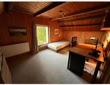 Holzvertäfeltes Zimmer mit schräger Decke, eingerichtet mit einem Schreibtisch, einem Stuhl, einer Tischlampe und einem Bett. Ein großes Fenster mit Blick ins Grüne lässt Tageslicht herein, und an der Wand hängt ein Landschaftsbild.