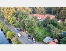 Luftaufnahme einer ländlichen Umgebung mit einem großen Haus mit rotem Satteldach, umgeben von Bäumen und Grünflächen. Eine schmale Straße mit parkenden Autos verläuft entlang eines Flusses, der links im Bild zu sehen ist.