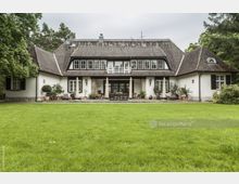 Großes, zweistöckiges Landhaus mit Satteldach, weißen Wänden und dunklen Fensterrahmen, umgeben von einem gepflegten, grünen Rasen und Bäumen. Vor dem Haus sind eine Terrasse mit Gartenmöbeln und zahlreiche Kübelpflanzen angeordnet.