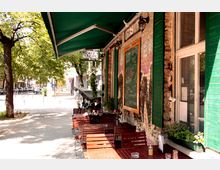 Außenbereich eines kleinen Cafés mit grünem Sonnendach, Holztischen und Stühlen auf einem Bürgersteig. Die Fassade besteht aus Backstein mit grünen Fensterläden, und eine mit Kreide beschriebene Tafel zeigt Speisen und Getränke. Bäume säumen die angrenzende Straße.