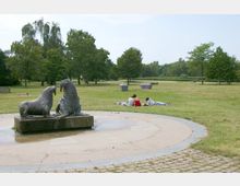 Ein Park mit einer Wiese, auf der eine Skulptur von zwei Walrossen auf einem Sockel im Vordergrund steht. Im Hintergrund sind Bäume, verstreut liegende Personen auf Decken und Müllbehälter zu sehen.