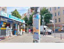 Straßenszene in einer städtischen Umgebung mit einem kleinen Kiosk namens „Eastside Kiosk“ im Vordergrund, der Waren wie Getränke und Zeitungen anbietet. Im Hintergrund sind Wohnhäuser, ein Drogeriemarkt, Bäume und ein Verkehrsschild mit der Beschriftung „Warschauer Straße“ zu sehen, sowie ein vorbeifahrender Bus und ein parkender Lieferwagen.