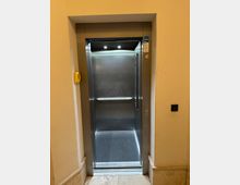 Offener Aufzug in einem Innenflur mit Edelstahlkabine, Deckenleuchten und seitlichem Bedienpanel. Der Aufzug ist in eine helle Wand eingelassen, mit Türrahmen aus Metall sowie Rufknopf und Hinweisschildern neben der Öffnung.