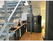 Moderne Küche mit offener Wendeltreppe aus Metall, einer schwarzen Kühl-Gefrierkombination, eingebauten Elektrogeräten und einer Arbeitsfläche aus Holz. Der Raum hat Holzboden, helle Wandfliesen und wird durch Tageslicht beleuchtet.