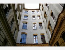 Blick in einen engen Innenhof eines mehrstöckigen Gebäudes mit hellen, verputzten Wänden, rot umrahmten Fenstern und Ziegeldekor an den Fassaden. Die Architektur zeigt historische Elemente mit Simsen und Ornamenten.