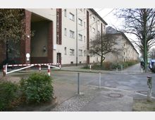 Wohnblock in einer urbanen Gegend mit hellbeigen Fassaden, roten Backsteinelementen und kleinen Fenstern. Im Vordergrund befinden sich eine Schranke, Gehwege mit Gehölzen sowie ein asphaltierter Bereich mit Pollern.