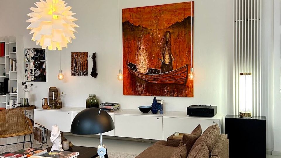 Modern living room with white walls and high ceiling beams, featuring a brown sectional sofa facing a low black coffee table on a light rug. The space includes a large orange-toned painting above sleek white cabinetry, pendant and floor lamps, built-in shelving, and a sculptural branch-like ceiling fixture.