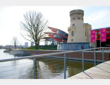 Uferansicht mit einem historischen Turm und einem modernen Gebäudekomplex mit roten, geschwungenen Vorsprüngen, umgeben von Wasser und Grünflächen. Im Vordergrund verläuft ein Geländer entlang des Wassers, und mehrere Personen spazieren auf der Promenade.