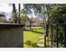 Ein gepflegter Garten mit grünem Rasen und vereinzelten Bäumen, aufgenommen durch ein schmiedeeisernes Tor. Im Hintergrund ist ein Haus mit rotem Ziegeldach und großen Fenstern sichtbar.