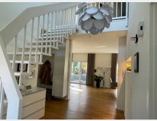 Moderne Wohnraumgestaltung mit einer weißen Treppe, die zu einem offenen Obergeschoss führt. Der Raum ist mit hellen Holzböden, großen Fenstern mit Vorhängen, einem dekorativen Kronleuchter und Pflanzenakzenten eingerichtet.