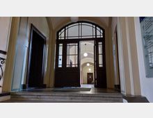 Innenansicht eines historischen Gebäudeeingangs mit hohen, hölzernen Flügeltüren und einem Bogenfenster darüber. Der dahinterliegende Korridor hat Gewölbedecken und ist durch mehrere runde Deckenlampen beleuchtet.