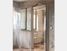 Ein minimalistisch eingerichteter Raum mit einer grauen Betonwand, einer offenen Glastür und einem Fenster im Hintergrund. Im angrenzenden Raum stehen zwei goldene Stühle mit gelben Sitzpolstern auf einem Holzboden, während ein durchscheinender Schleierstoff vor der Tür hängt.