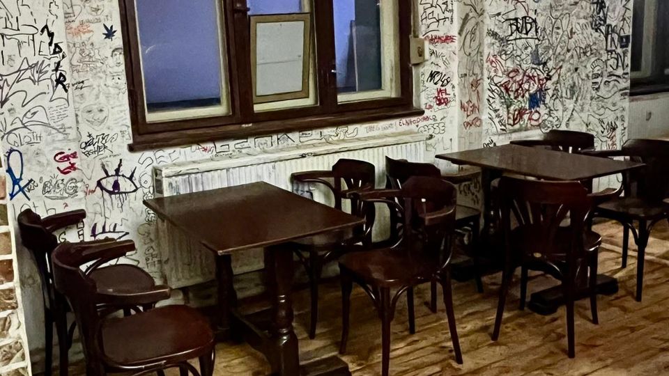 Innenraum eines einfachen Cafés oder Lokals mit Holzdielenboden, mehreren dunklen Holztischen und Stühlen sowie einem großen Holzfenster über einem Heizkörper. Die Wände sind bis zur halben Höhe weiß gestrichen und dicht mit Graffiti, Zeichnungen und Schriftzügen bedeckt.